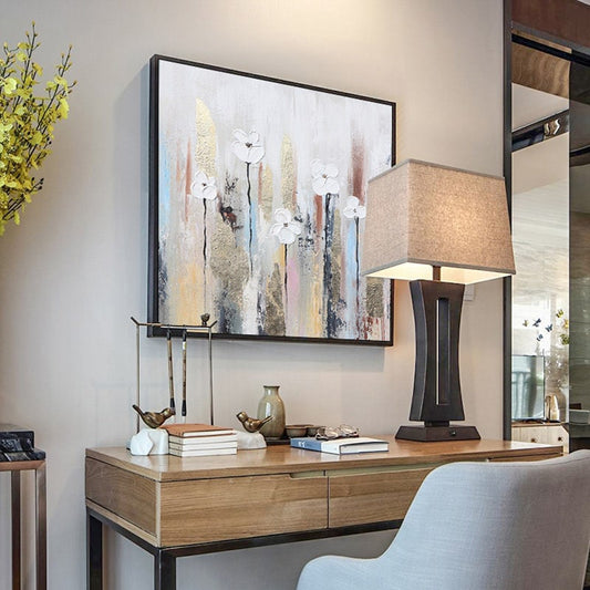 May include: A wooden desk with a light brown top and black metal legs. A lamp with a beige fabric shade sits on the desk. A painting with white flowers on a colorful background hangs on the wall above the desk.