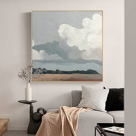 May include: An abstract landscape painting featuring a cloudy sky and a distant horizon. The artwork is framed in a gold-colored frame and hangs above a gray sofa. The painting's color palette includes shades of gray, white, brown, and blue.