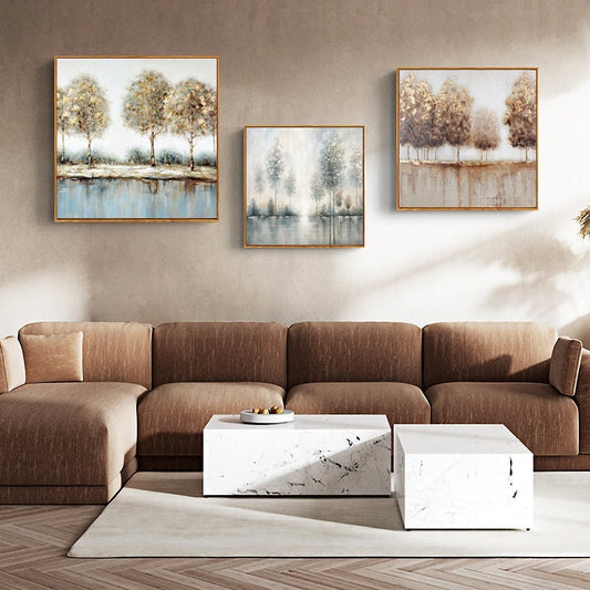 May include: Three abstract landscape paintings with a muted color palette of brown, gray, and blue. The paintings depict trees and a body of water. The paintings are framed in gold and hung on a beige wall above a brown sectional sofa.