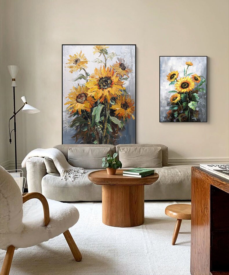 May include: Two framed paintings of sunflowers in a living room setting. The larger painting features a bouquet of sunflowers in shades of yellow and green against a gray background. The smaller painting features a smaller bouquet of sunflowers in a similar style.