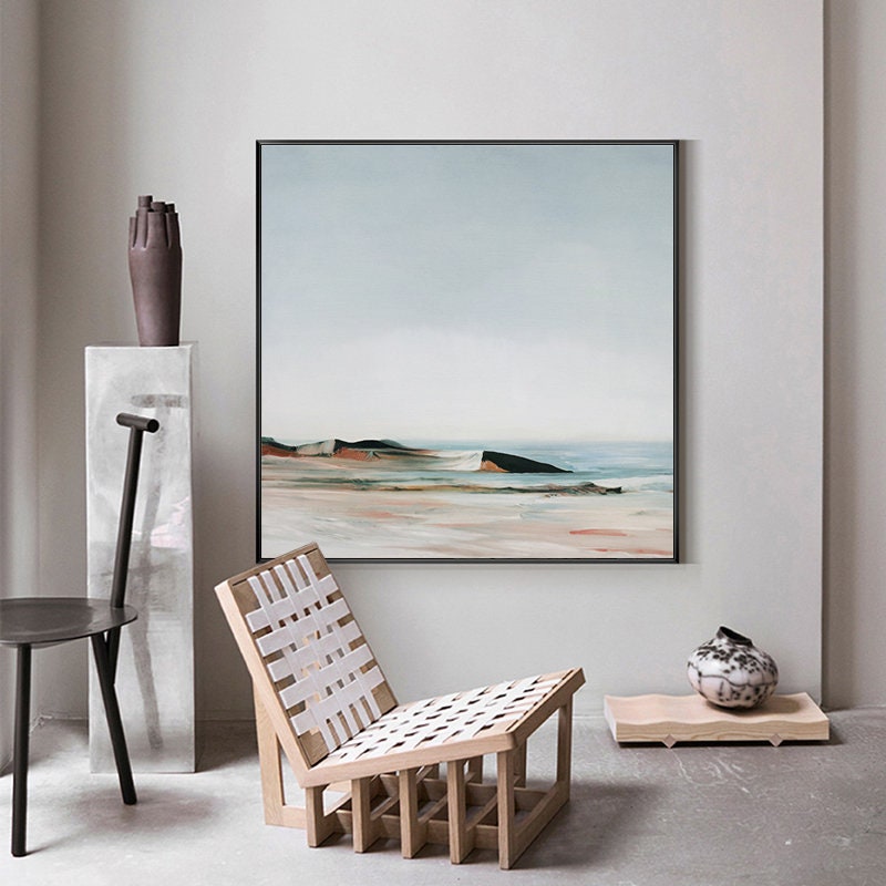 May include: A framed abstract painting of a seascape with a light blue sky, white clouds, and a sandy beach with a rocky shoreline. The painting is in a black frame and is hanging on a white wall.
