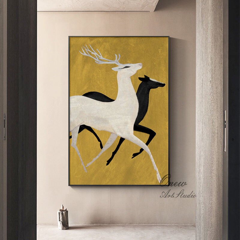 May include: Abstract painting of two deer running on a gold background. The deer in the foreground is white, while the deer in the background is black. The painting is framed in black.