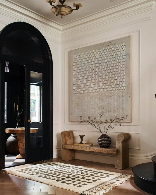 May include: A white rug with a geometric pattern is placed in front of a black door with a decorative arch. A stone bench with rounded ends sits against a white wall with a large abstract painting above it. A vase with dried branches sits on the bench.