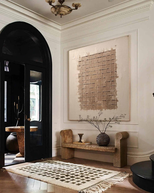 May include: A large abstract painting with a grid pattern in shades of brown and beige hangs on a white wall above a stone bench. The bench has a vase with dried branches and a small ceramic vase on top. The room has a black door with a white and black checkered floor.