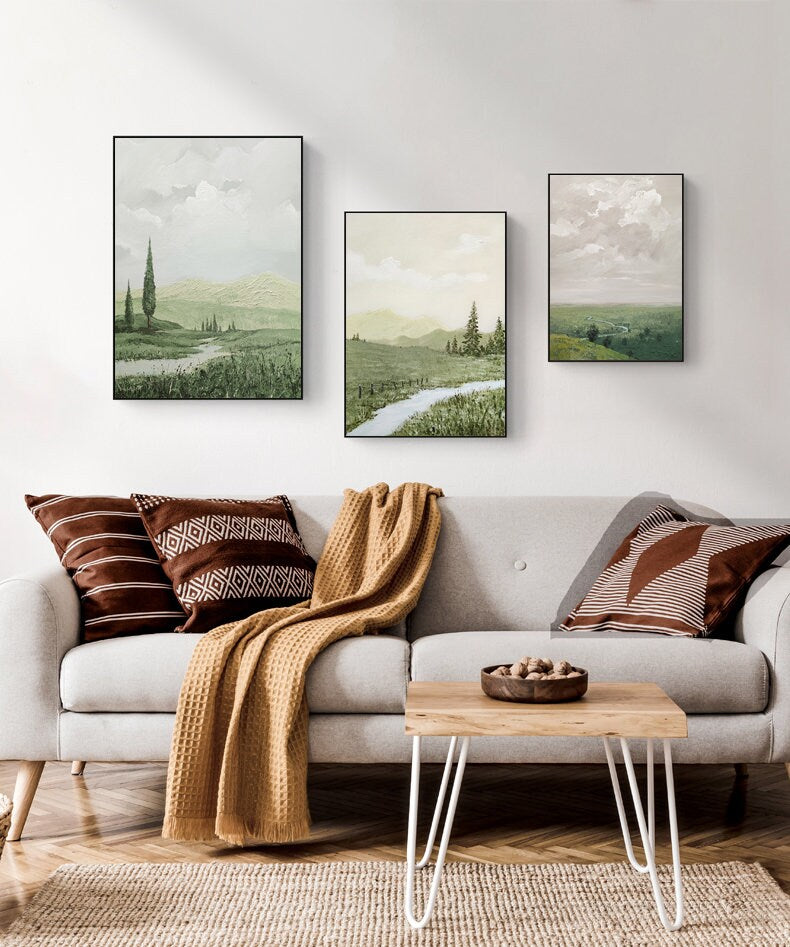 May include: Three framed landscape paintings with green and brown tones depicting rolling hills, trees, and a winding path. The paintings are hung on a white wall above a light gray sofa with brown and white patterned throw pillows and a brown throw blanket.