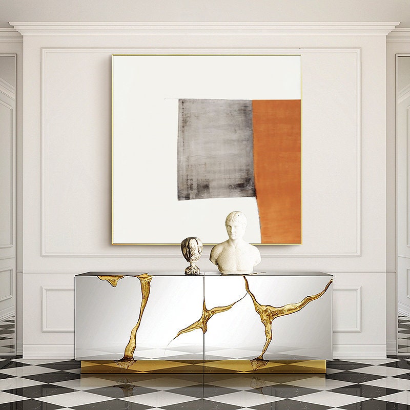 May include: A white wall with a gold framed abstract painting above a silver and gold mirrored console table. The console table has two doors and a gold base.