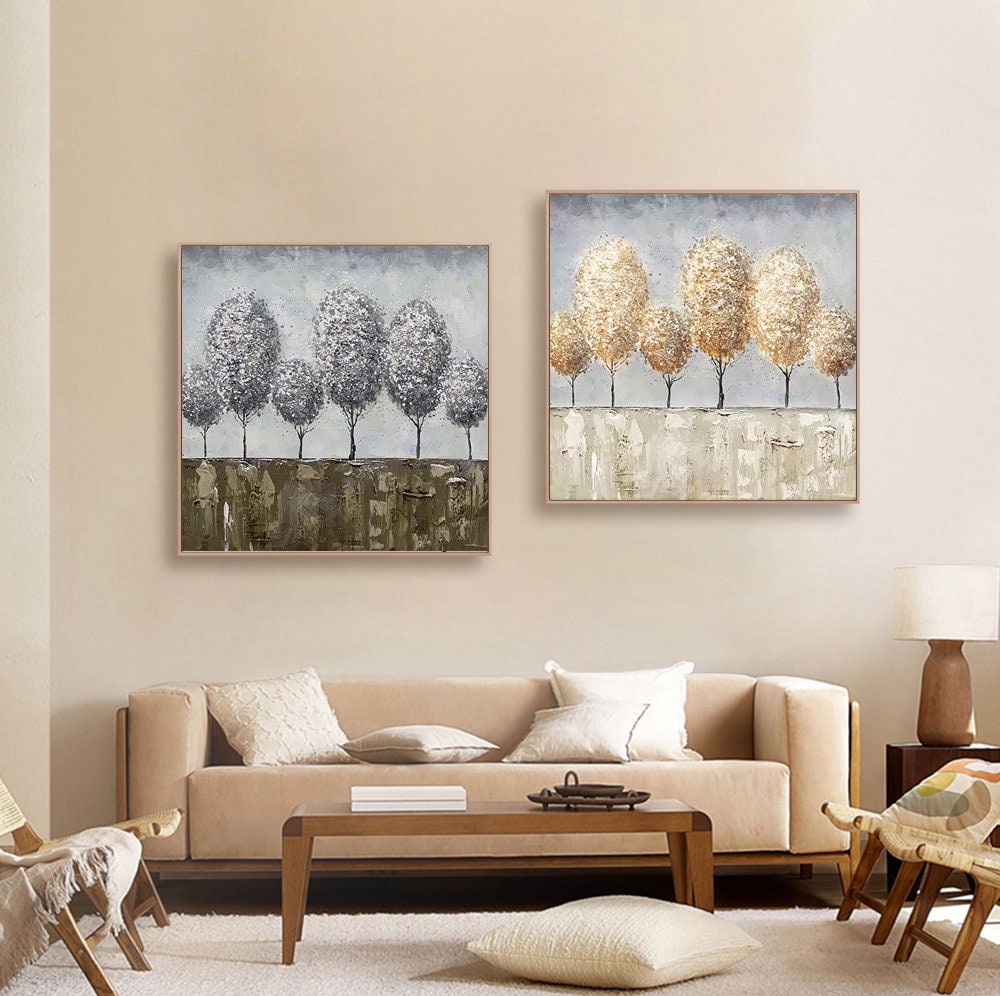 May include: Two framed abstract paintings of trees with a light gray background. The painting on the left features gray trees with brown and gold accents. The painting on the right features gold trees with white accents.