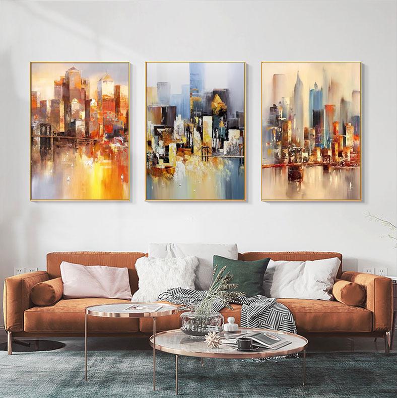 May include: Three abstract cityscape paintings with a muted color palette of blues, yellows, and oranges. The paintings are framed in gold and hung on a white wall.
