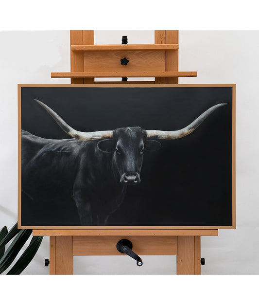 May include: A framed artwork featuring a black longhorn bull against a dark background. The bull's black coat contrasts with its large, light-colored horns. The artwork is displayed on a wooden easel.