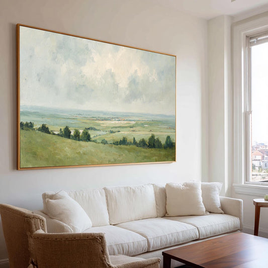 May include: A large landscape painting with a gold frame depicts a serene countryside scene. The artwork features rolling green hills, trees, and a cloudy sky with soft, muted colors. The painting hangs above a white sofa in a well-lit living room.