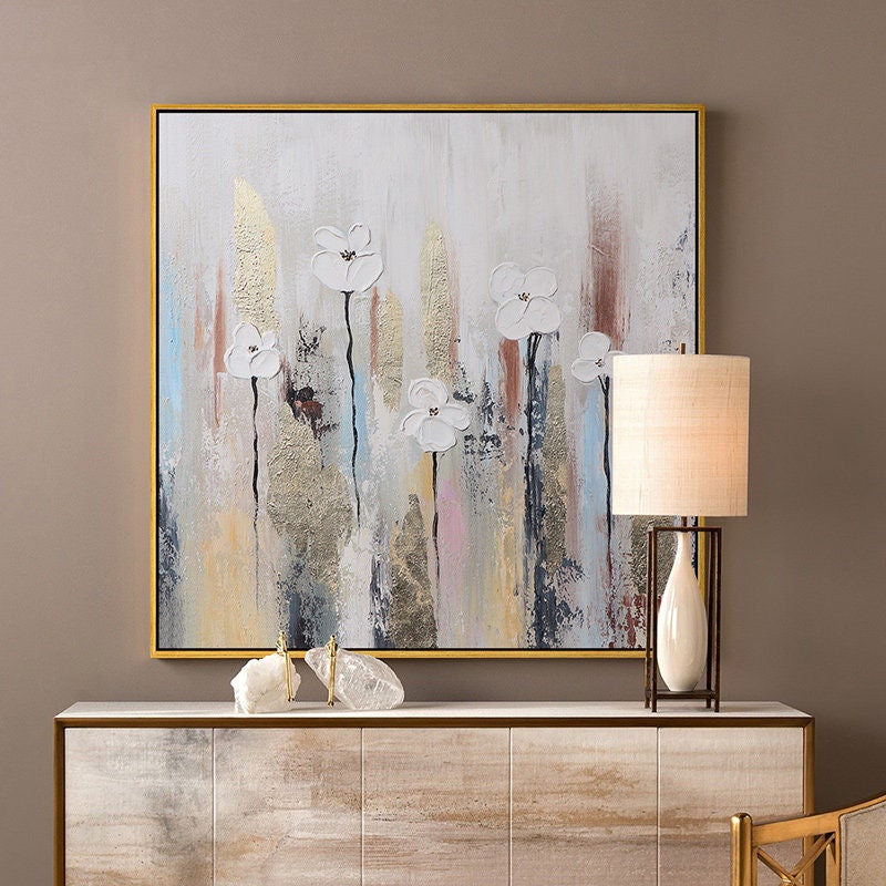 May include: Abstract painting with white flowers on a textured background with gold accents. The painting is framed in gold and is hanging on a wall above a wooden dresser.