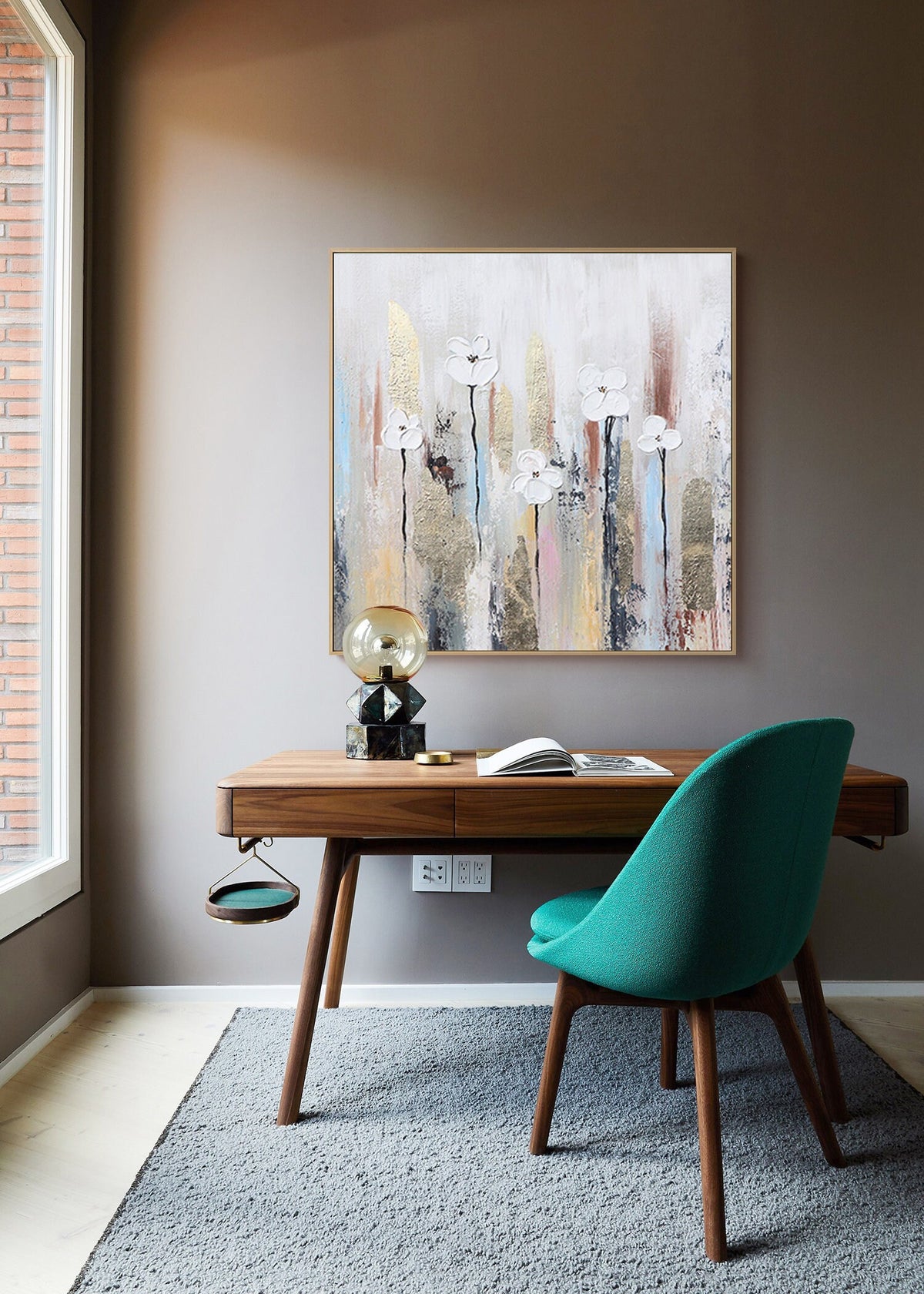 May include: A modern home office with a wooden desk and a teal chair. A painting with white flowers and gold accents hangs on the wall above the desk. A lamp with a clear glass globe sits on the desk.