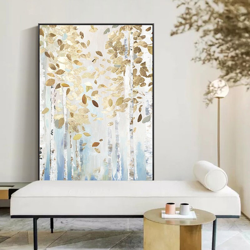 May include: Abstract art print featuring a forest scene with gold and brown leaf accents. The painting has a white, blue, and gold color palette. The artwork is framed in a black rectangular frame. The painting is displayed in a modern living room setting.