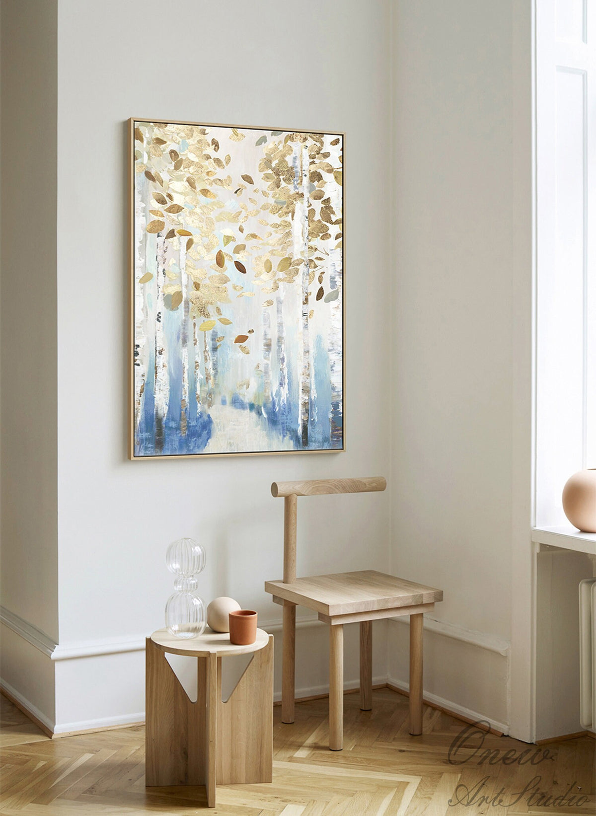 May include: Abstract forest painting with gold and brown leaves, blue and white trees, and a gold frame. The artwork hangs on a white wall above a small wooden table and chair.