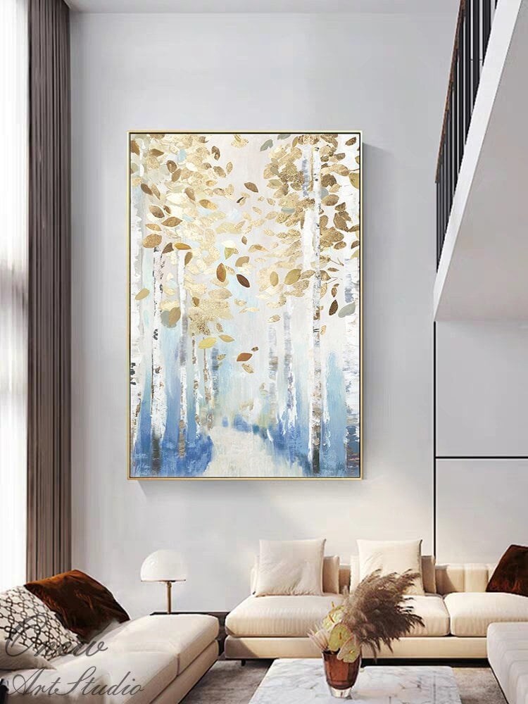 May include: Large framed abstract painting featuring a forest scene with gold and brown leaves. The artwork has a gold frame and a color palette of white, blue, and gold. The painting is displayed in a modern living room setting.