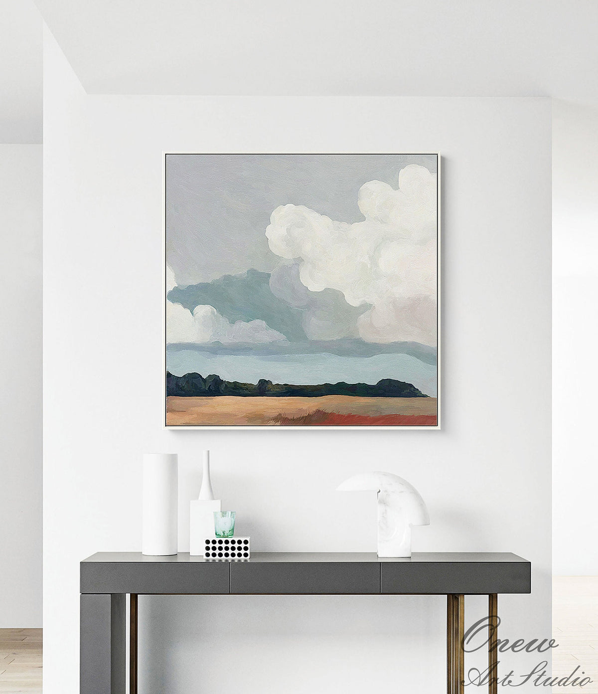 May include: Abstract landscape painting with a silver frame. The artwork depicts a cloudy sky above a field. The color palette includes shades of blue, white, brown, and green. The painting is displayed on a dark gray console table with decorative items.