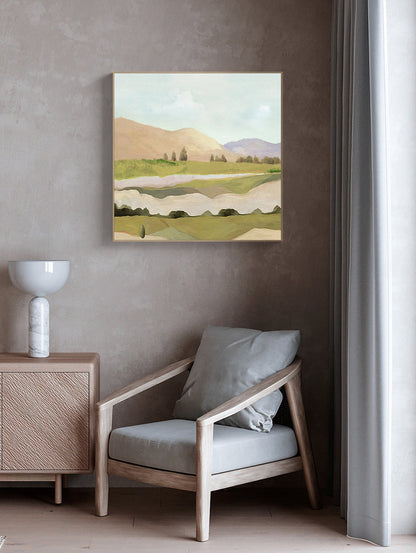 May include: A framed abstract landscape painting with a light blue sky, green hills, and a brown river. The painting is hanging on a wall in a room with a light brown armchair and a wooden side table.