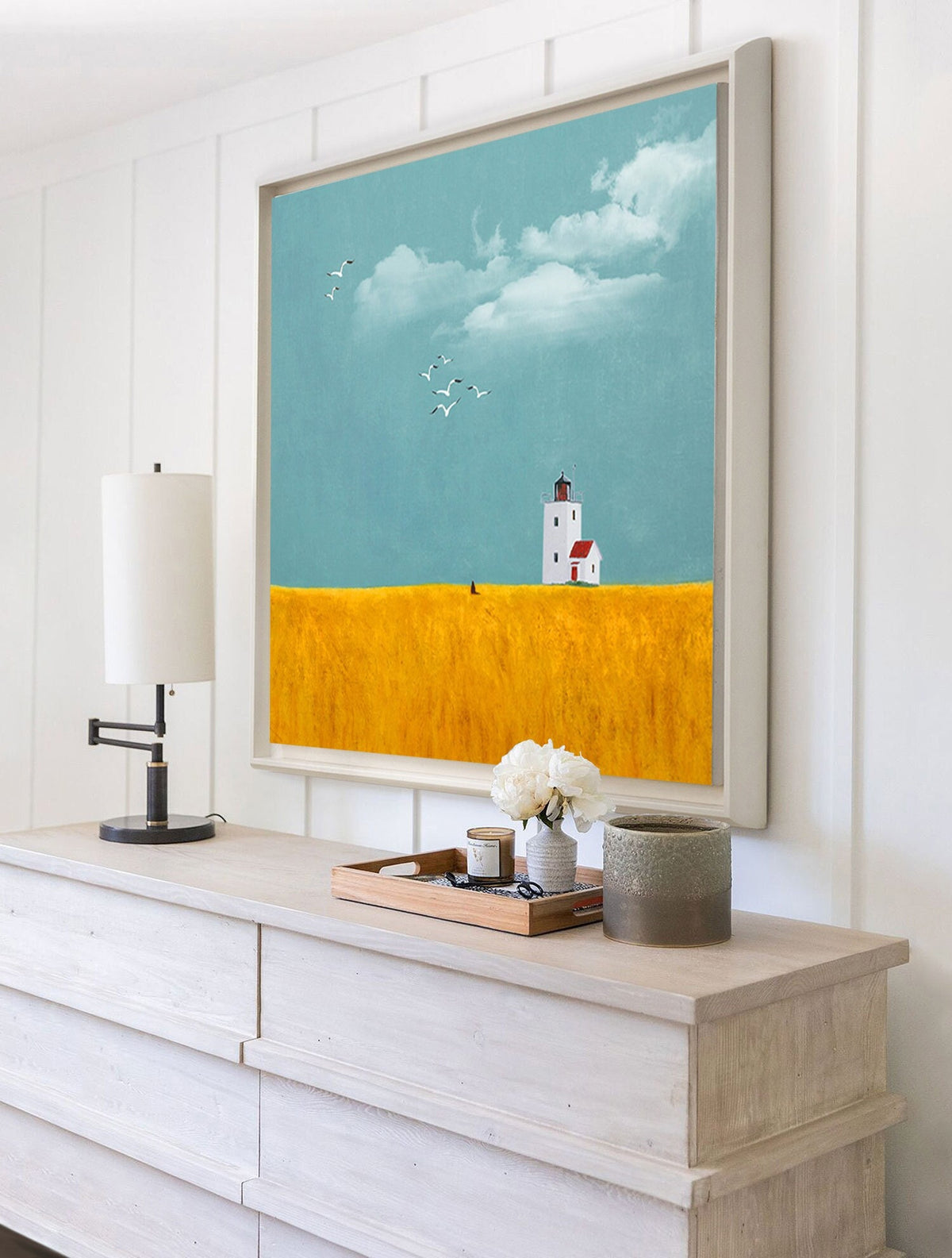 May include: A framed painting of a white lighthouse with a red roof on a yellow field with a blue sky and white clouds. There are birds flying in the sky. The painting is hanging on a white wall above a white dresser with a lamp and a tray of candles and flowers.