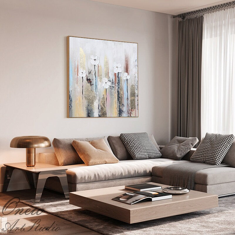 May include: A modern living room with a white couch and a large abstract painting of white flowers on a beige background. The painting is framed in gold and hangs above the couch. The couch has a variety of pillows in different colors and patterns. A coffee table sits in front of the couch with a book on top. The room is decorated in a neutral color palette with a gold lamp on a side table.