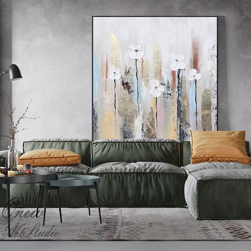 May include: Abstract painting with white flowers and gold accents on a canvas. The background is a mix of light and dark colors, including blue, pink, and brown.
