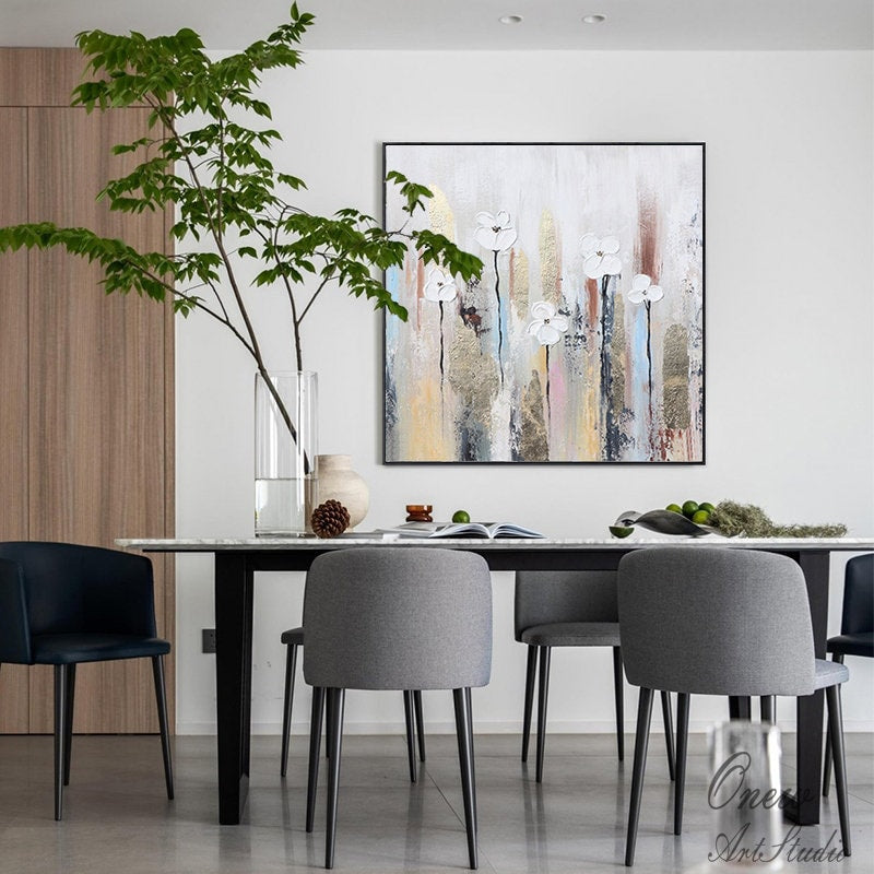 May include: A modern dining room with a white and gold abstract painting featuring white flowers on a textured background. The dining table has a white marble top and black legs. There are four chairs around the table, two gray and two black. A large glass vase with green branches is on the table.