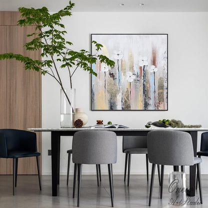May include: A modern dining room with a white and gold abstract painting featuring white flowers on a textured background. The dining table has a white marble top and black legs. There are four chairs around the table, two gray and two black. A large glass vase with green branches is on the table.