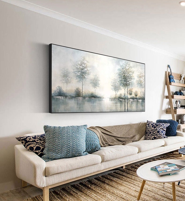 May include: A large rectangular landscape painting with a black frame depicts a serene waterscape with trees. The artwork hangs above a cream-colored sofa adorned with blue and patterned throw pillows and a tan blanket. A round white coffee table and a woven rug complete the living room setting.
