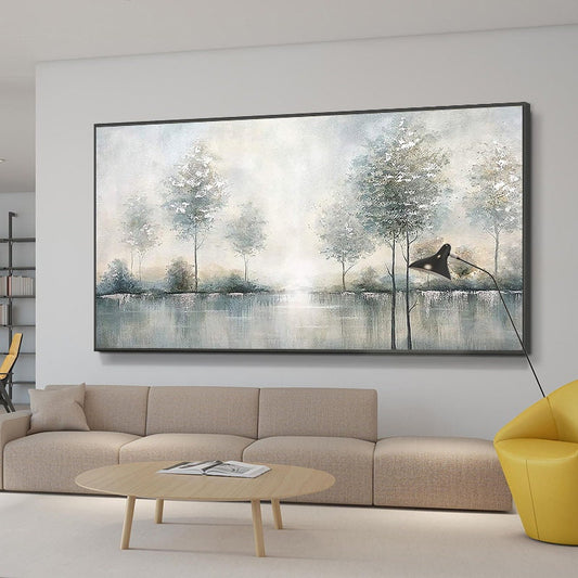 May include: Large landscape painting featuring a serene waterscape with trees in muted tones of gray, white, and blue. The artwork is framed in a black border and hangs above a beige sofa. A modern black lamp and yellow chair are also in the room.