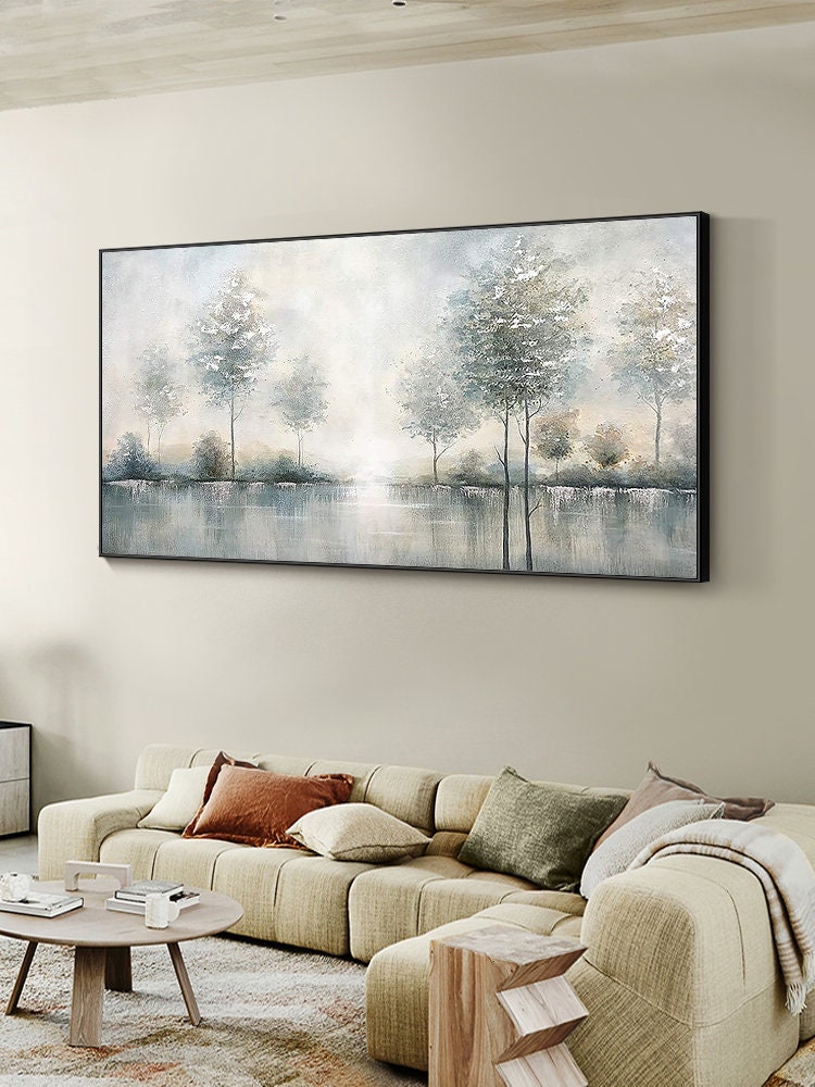 May include: A large rectangular landscape painting in a black frame. The artwork depicts a serene waterscape with trees along the water's edge. The color palette is soft, with muted blues, greens, and whites, creating a tranquil atmosphere. The painting hangs above a beige sectional sofa.