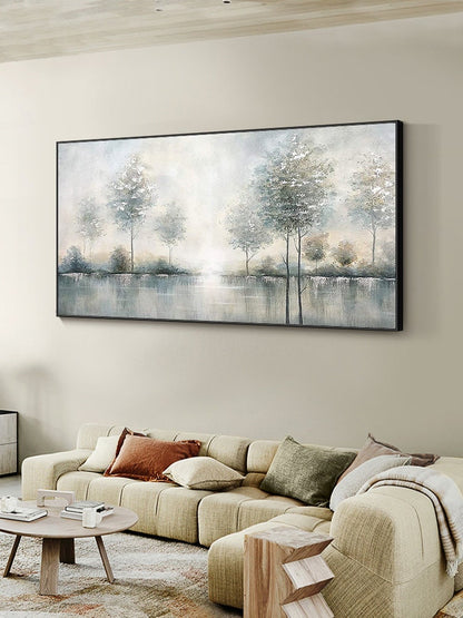 May include: A large rectangular landscape painting in a black frame. The artwork depicts a serene waterscape with trees along the water's edge. The color palette is soft, with muted blues, greens, and whites, creating a tranquil atmosphere. The painting hangs above a beige sectional sofa.