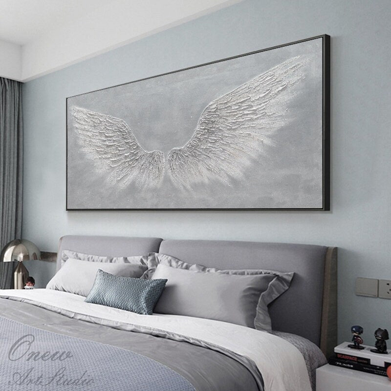 May include: A large, rectangular canvas painting with a gray background and white, textured angel wings. The painting is hanging on a wall above a gray headboard and bed.