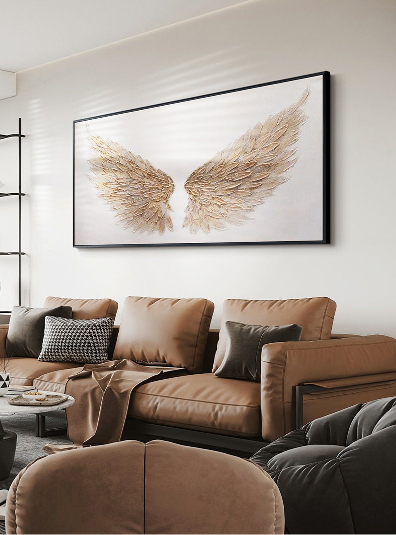 May include: A framed artwork featuring a pair of textured, golden angel wings against a white background. The artwork is displayed above a brown leather sofa with various throw pillows. The room has a modern, minimalist design.