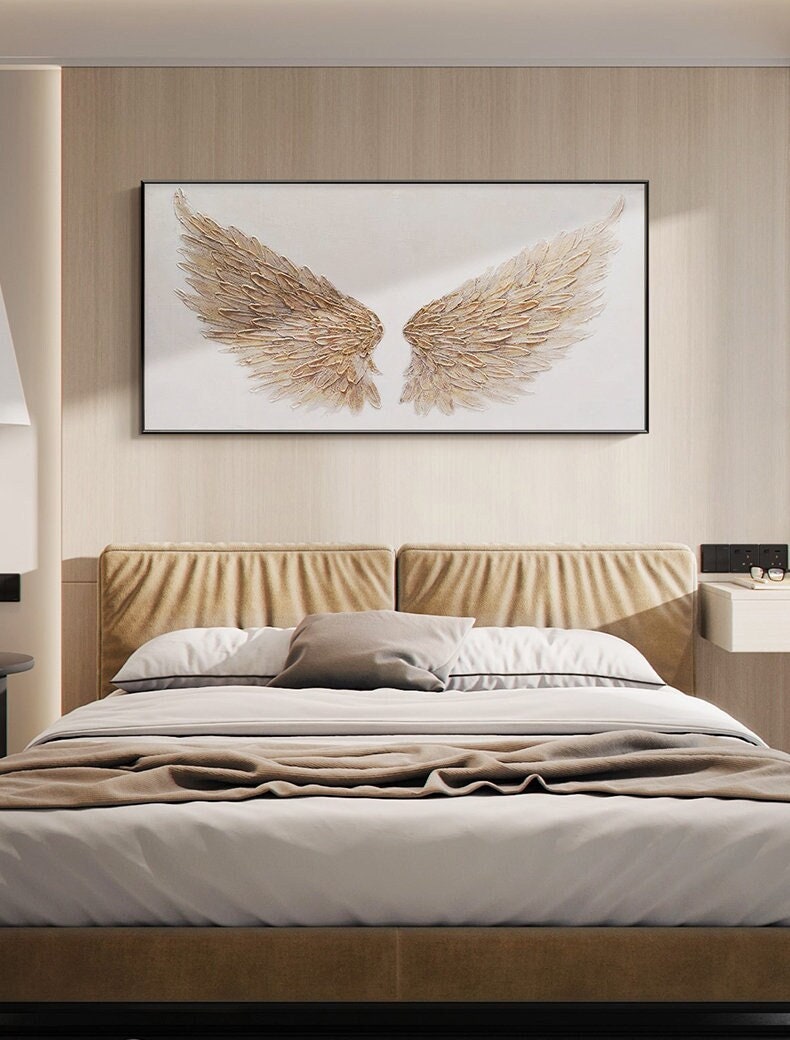 May include: A framed artwork featuring a pair of gold-colored angel wings against a white background. The wings have a textured, feathered appearance. The artwork hangs above a bed with a tan headboard, white bedding, and a brown blanket.