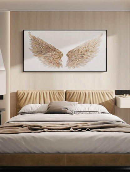 May include: A framed artwork featuring a pair of gold-colored angel wings against a white background. The wings have a textured, feathered appearance. The artwork hangs above a bed with a tan headboard, white bedding, and a brown blanket.