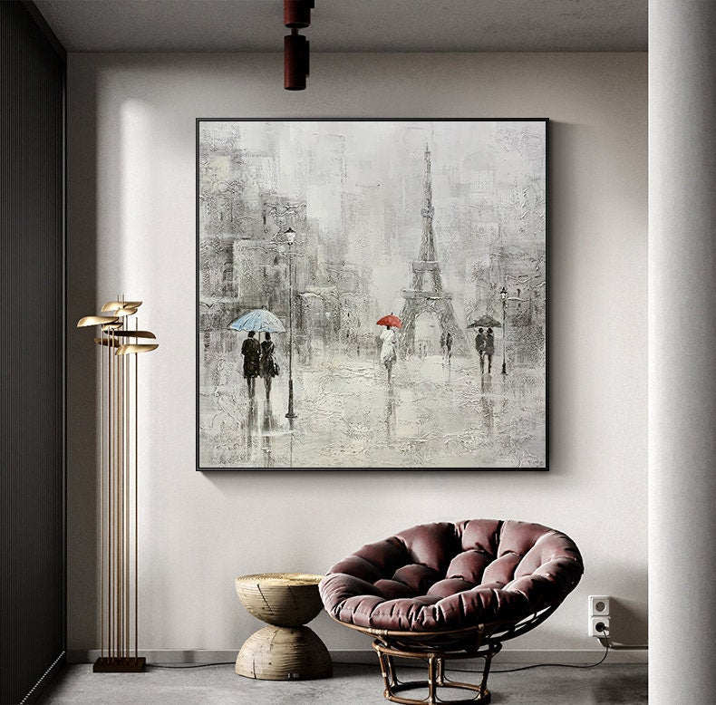 May include: A painting of a Parisian street scene with the Eiffel Tower in the background. The painting is done in a gray and white color scheme with pops of red and blue from the umbrellas. The painting is framed and hanging on a wall.