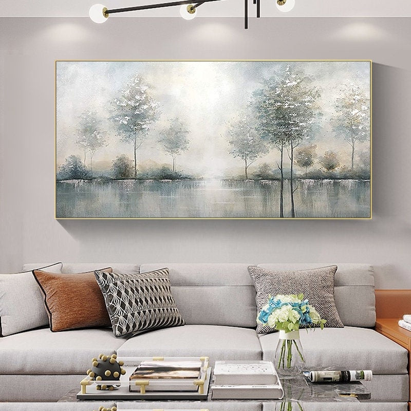 May include: A rectangular landscape painting with a gold frame depicts a serene scene of trees reflected in a body of water. The artwork uses soft, muted tones of gray, blue, and white, creating a tranquil atmosphere. The painting is displayed above a gray sofa with decorative pillows.