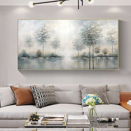 May include: A rectangular landscape painting with a gold frame depicts a serene scene of trees reflected in a body of water. The artwork uses soft, muted tones of gray, blue, and white, creating a tranquil atmosphere. The painting is displayed above a gray sofa with decorative pillows.