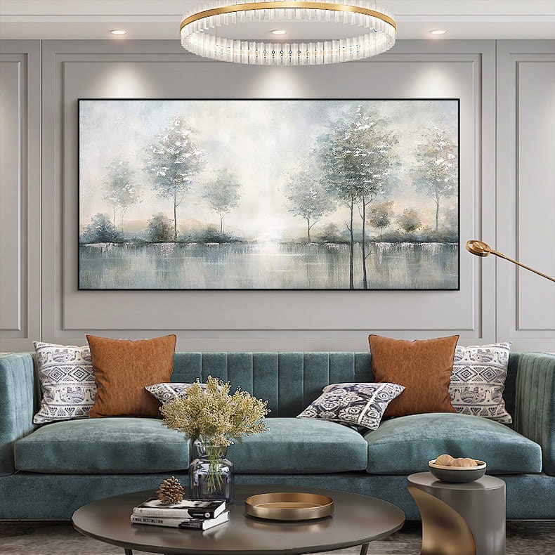 May include: A large rectangular landscape painting with a black frame. The artwork depicts a serene scene of trees reflected in a body of water, with soft, muted colors of gray, white, and blue. The painting hangs above a teal sofa with decorative pillows.