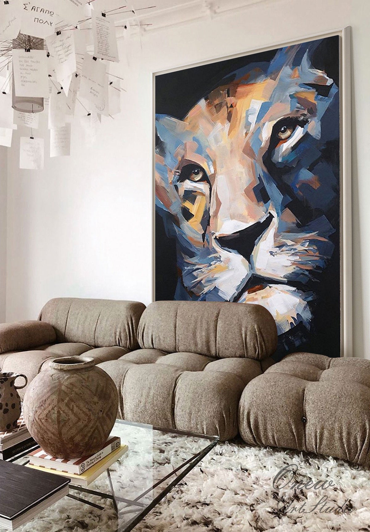 May include: Large framed abstract painting of a lion's face in shades of blue, gold, and white. The artwork is displayed above a brown sectional sofa and a glass coffee table. The room has a modern, minimalist design.