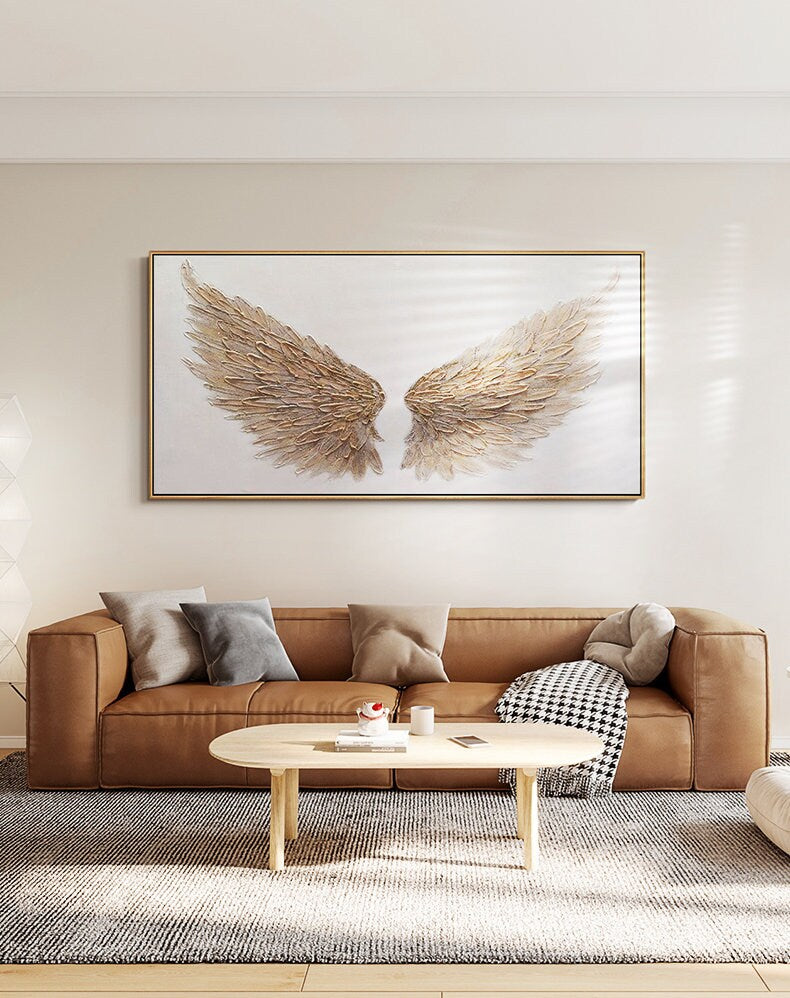 May include: A large rectangular framed canvas art piece featuring a pair of stylized angel wings. The wings are rendered in shades of gold and brown against a white background. The artwork is displayed above a brown leather sofa.