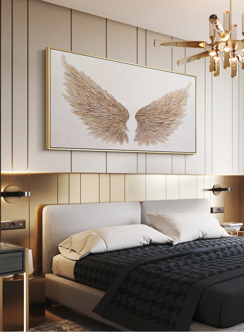 May include: A framed artwork featuring a pair of golden angel wings against a white background. The wings have a textured, feathered appearance and are set within a gold frame. The artwork hangs above a bed with a gray headboard and black and white patterned bedding.