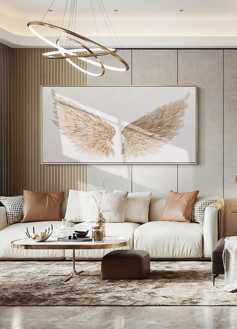 May include: A large, rectangular artwork featuring a pair of golden angel wings against a white background. The wings are textured and detailed, with a gold frame. The artwork hangs above a white sofa with brown and white pillows.