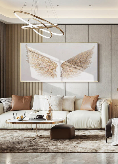 May include: A large, rectangular artwork featuring a pair of golden angel wings against a white background. The wings are textured and detailed, with a gold frame. The artwork hangs above a white sofa with brown and white pillows.