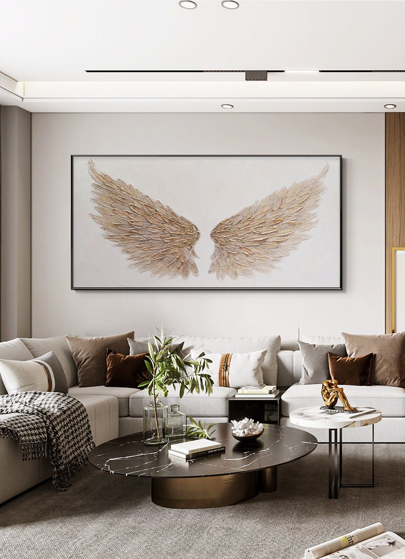 May include: A large, rectangular wall art piece featuring a pair of textured, golden-brown angel wings against a white background. The artwork is framed in black and hangs above a modern sofa in a living room setting.