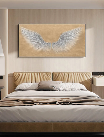 May include: A white canvas painting of a pair of angel wings on a beige background. The painting is framed in black and hanging above a bed with a tan headboard.