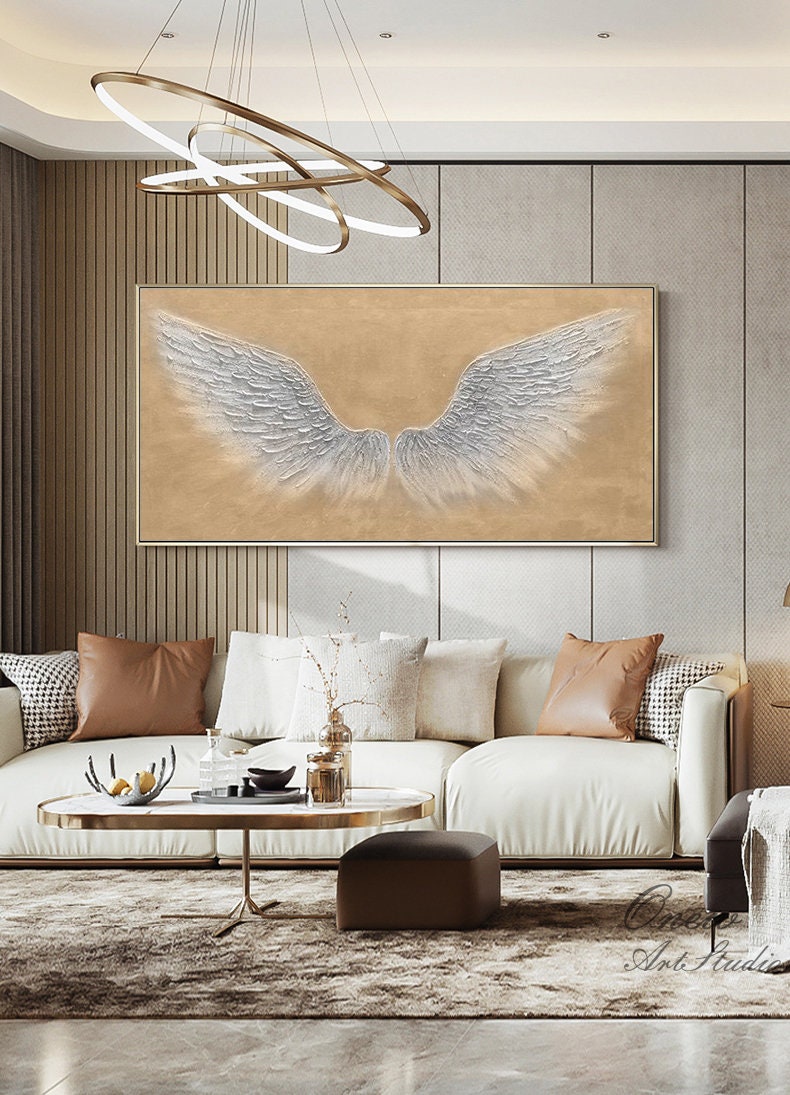 May include: A white canvas painting with a large, silver-colored pair of angel wings on a beige background. The painting is hanging on a wall in a living room with a white sofa and a coffee table.