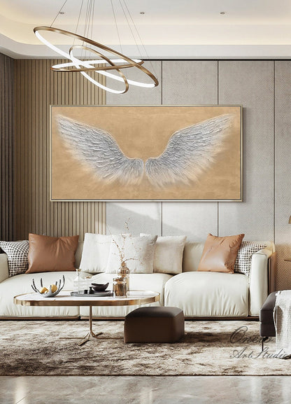 May include: A white canvas painting with a large, silver-colored pair of angel wings on a beige background. The painting is hanging on a wall in a living room with a white sofa and a coffee table.