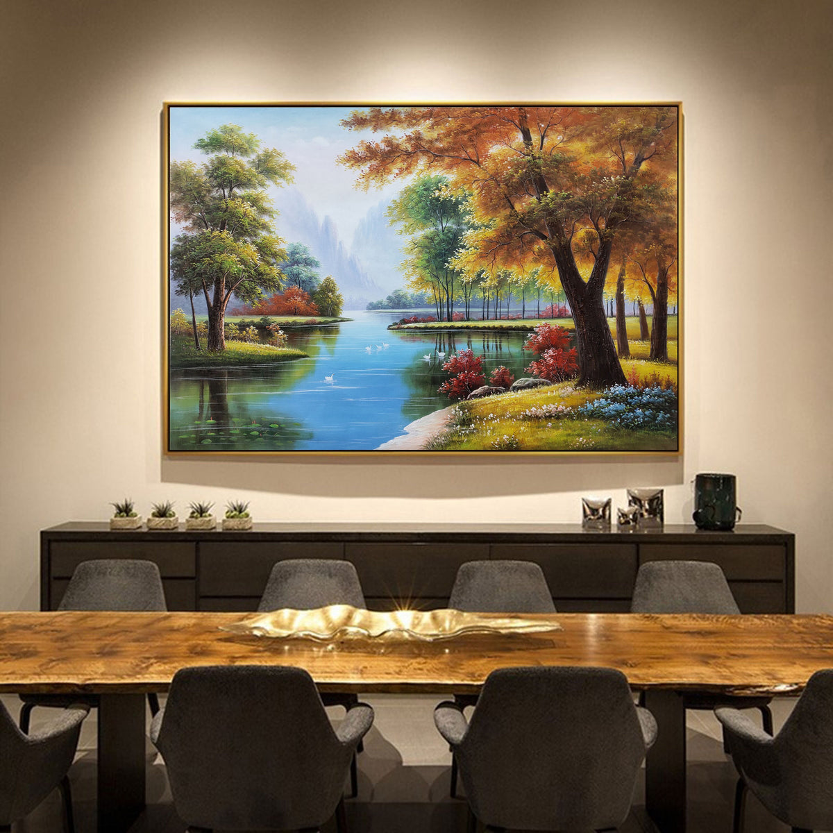 May include: A framed landscape painting depicting a river flowing through a forest with colorful trees and a blue sky. The painting is hanging on a wall above a wooden table with chairs.