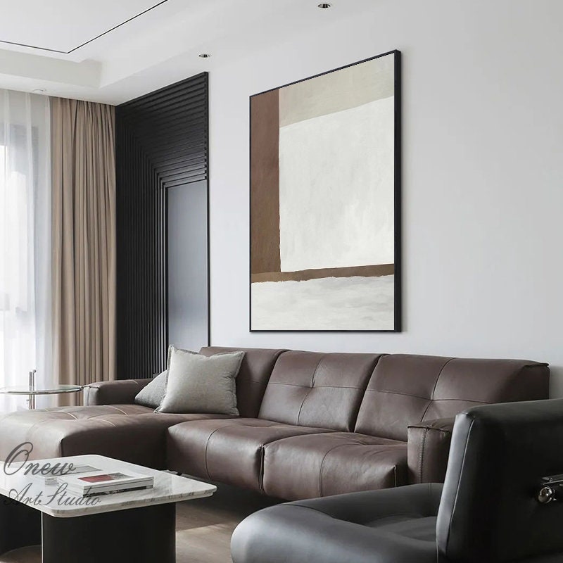 May include: A brown and white abstract painting in a black frame hangs on a white wall above a brown leather sectional sofa.