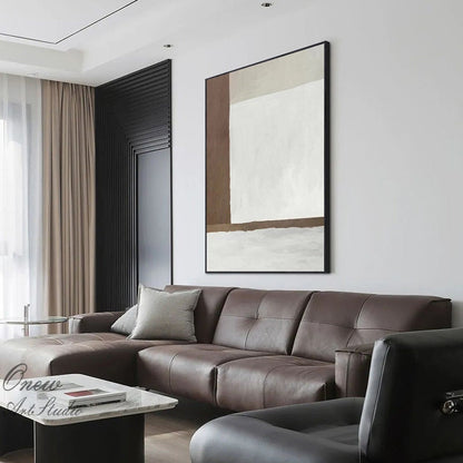 May include: A brown and white abstract painting in a black frame hangs on a white wall above a brown leather sectional sofa.